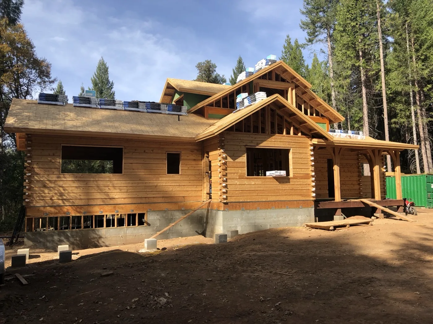 Log home shell with its roof sheathed and shingle bundles staged along the ridges, porch posts set at the right, at the edge of a pine forest