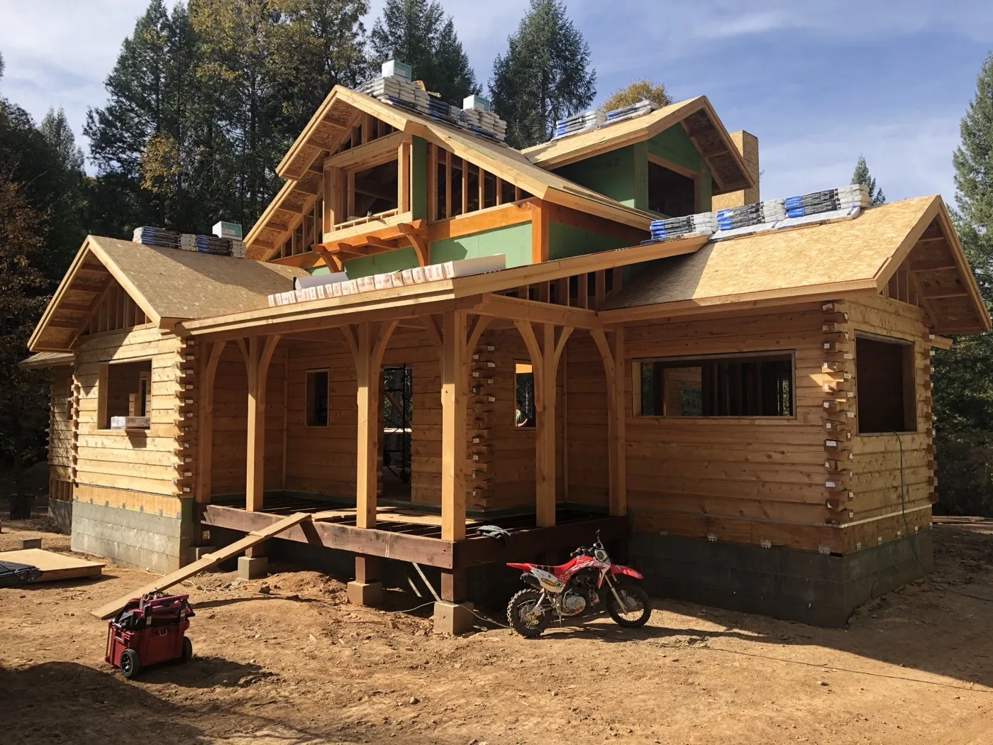 Log home under construction with a timber-framed porch across the front, shingle bundles staged on the sheathed roofs, and a dirt bike parked at the deck framing