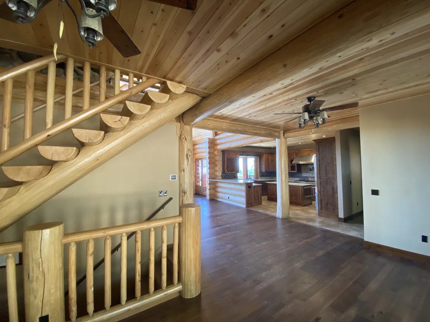 Finished interior: a log staircase with half-log treads and log railings, wood-clad ceilings with fans, and the kitchen beyond the log posts