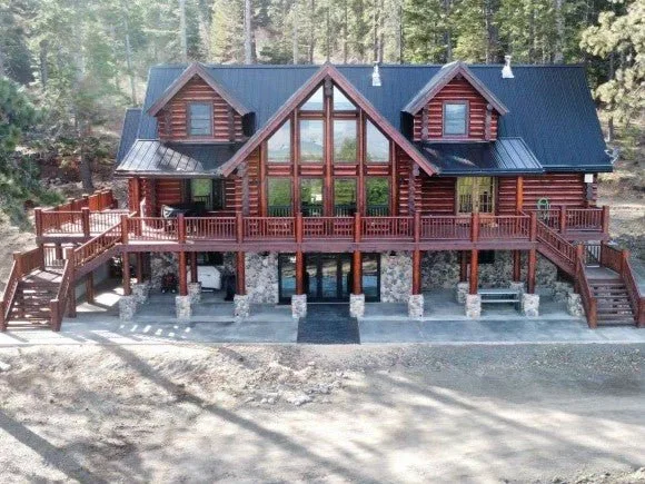Finished log home from the air: a full-width deck on stone piers, a two-story wall of windows under the central gable, and dark metal roofs against the hillside pines