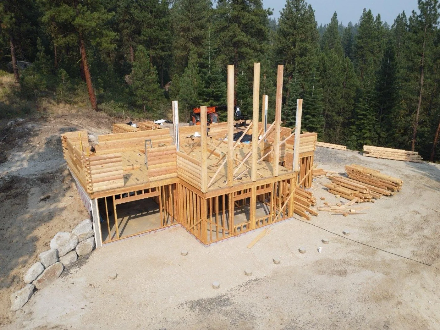 Build site from the air: log walls and tall posts rising over a framed walk-out level, with stacks of milled logs waiting on the pad among the pines
