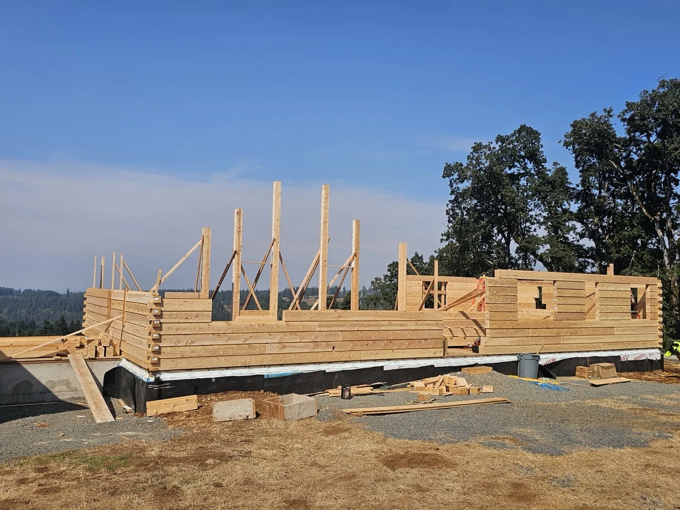 Log walls at window height with braced posts against a clear sky, on a hilltop pad above a wooded valley